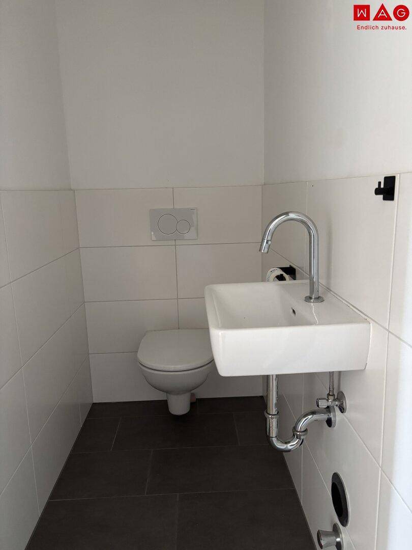 Separates WC mit wandhängender Toilette und kleinem Handwaschbecken in weißer Fliesenoptik.