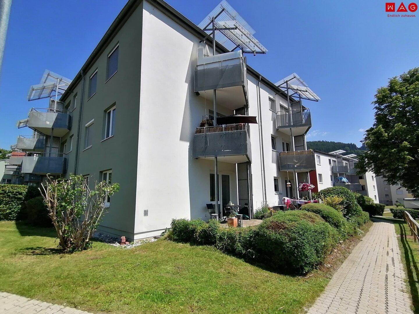 Modernes Mehrfamilienhaus mit Balkonen, einer Terrasse im Erdgeschoss und gepflegtem Garten unter blauem Himmel.