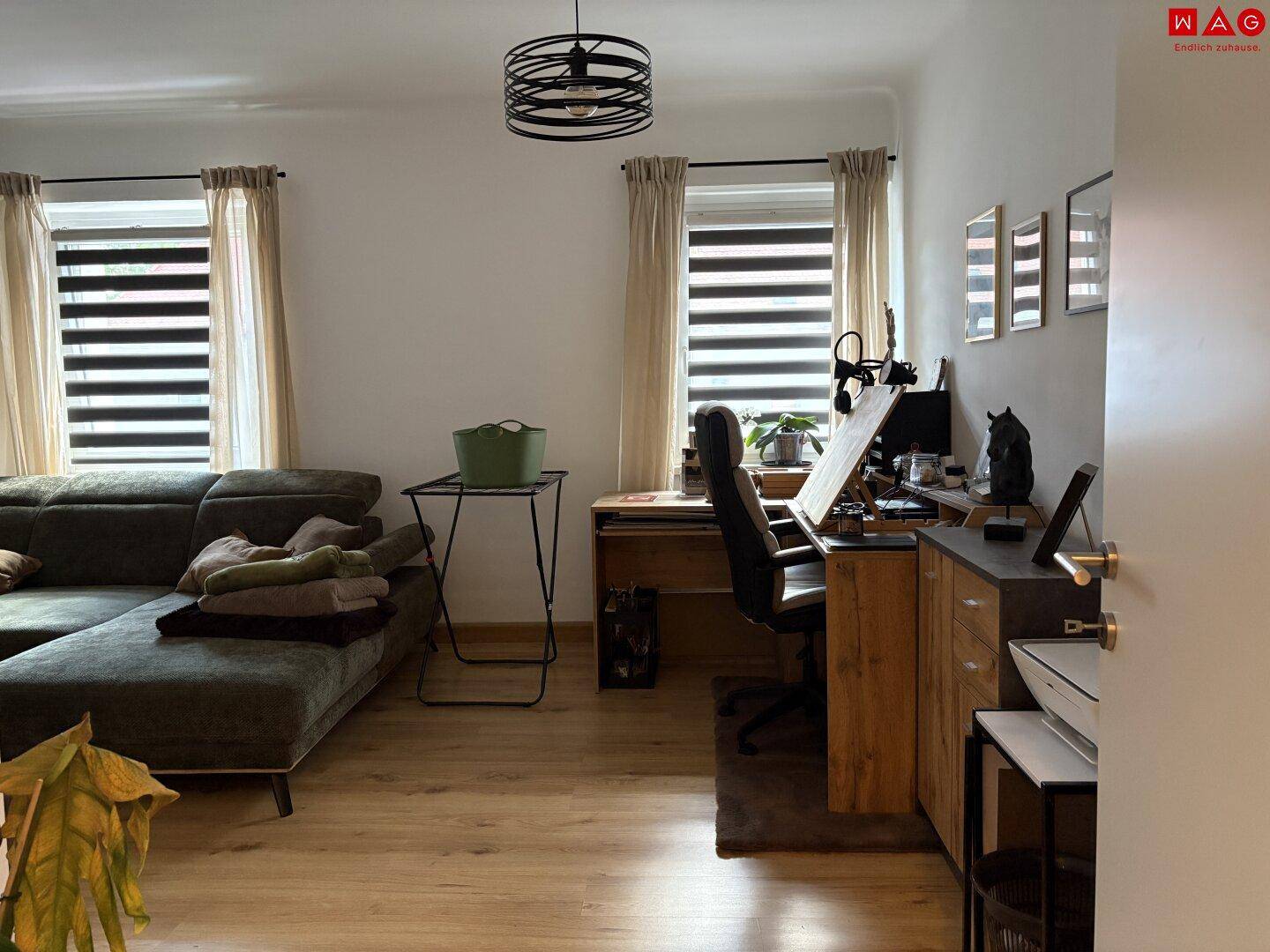 Geräumiges Wohnzimmer mit Ecksofa, Arbeitsbereich und großen Fenstern für viel Tageslicht.