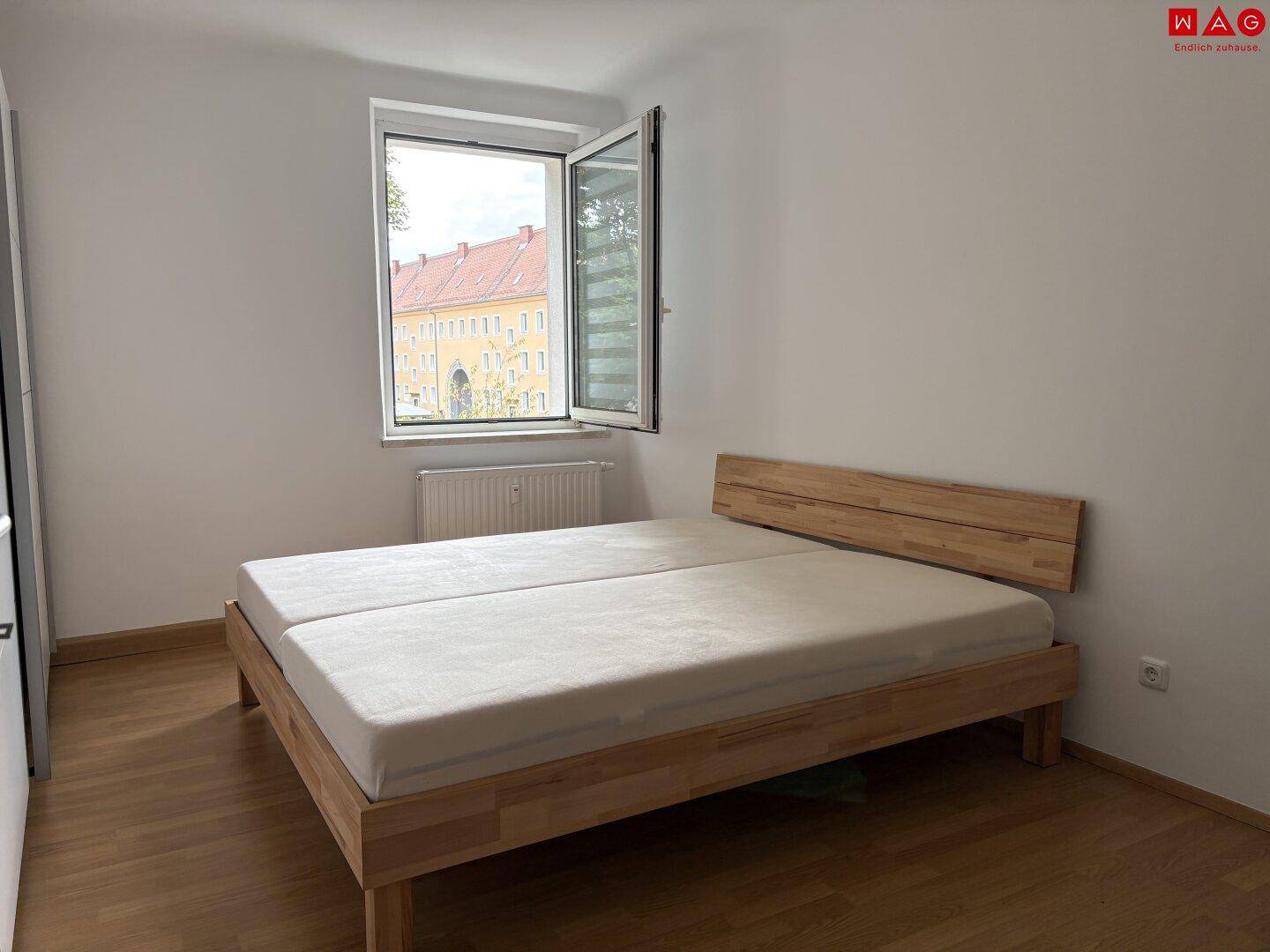 Helles Schlafzimmer mit Holzbettrahmen, Parkettboden und Fenster mit Blick auf die Umgebung.