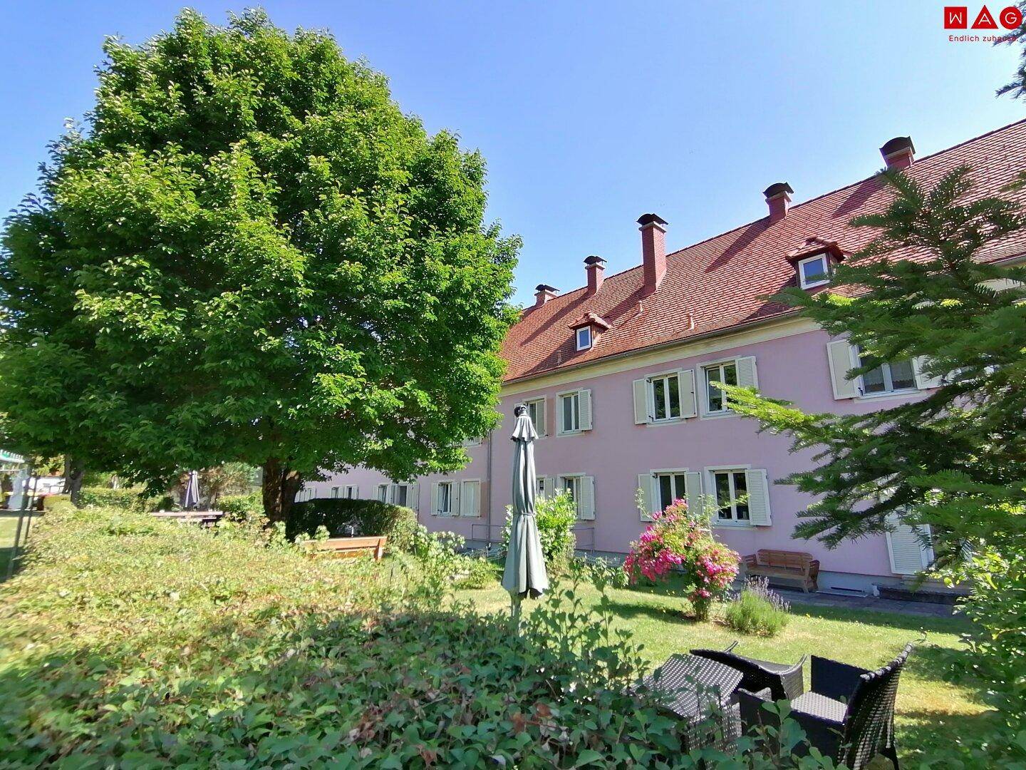 Gepflegter Gartenbereich mit Sitzgelegenheiten und altem Baumbestand vor einem traditionellen Gebäude.