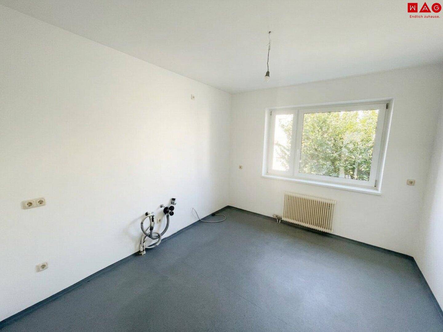 Leere Küche mit dunklem Boden, Fenster und vorbereiteten Anschlüssen für Küchengeräte.