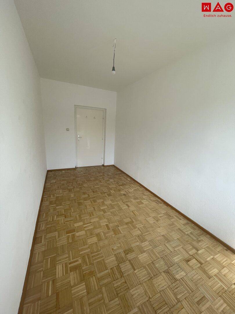 Leeres Zimmer mit klassischem Parkettboden und schlichten, weißen Wänden.