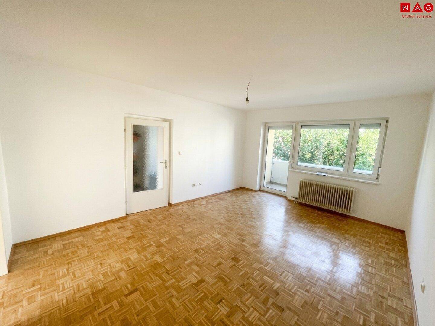 Geräumiges Wohnzimmer mit Fischgrätparkett, hellen Wänden und Zugang zum Balkon.
