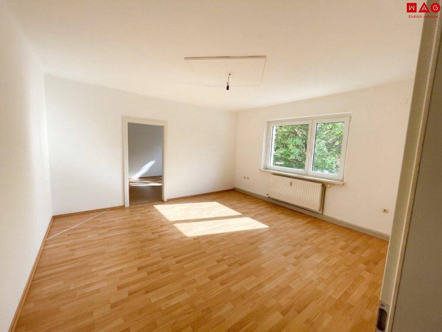 Helles, leeres Wohnzimmer mit Parkettboden, weiß gestrichenen Wänden und Blick ins Grüne.