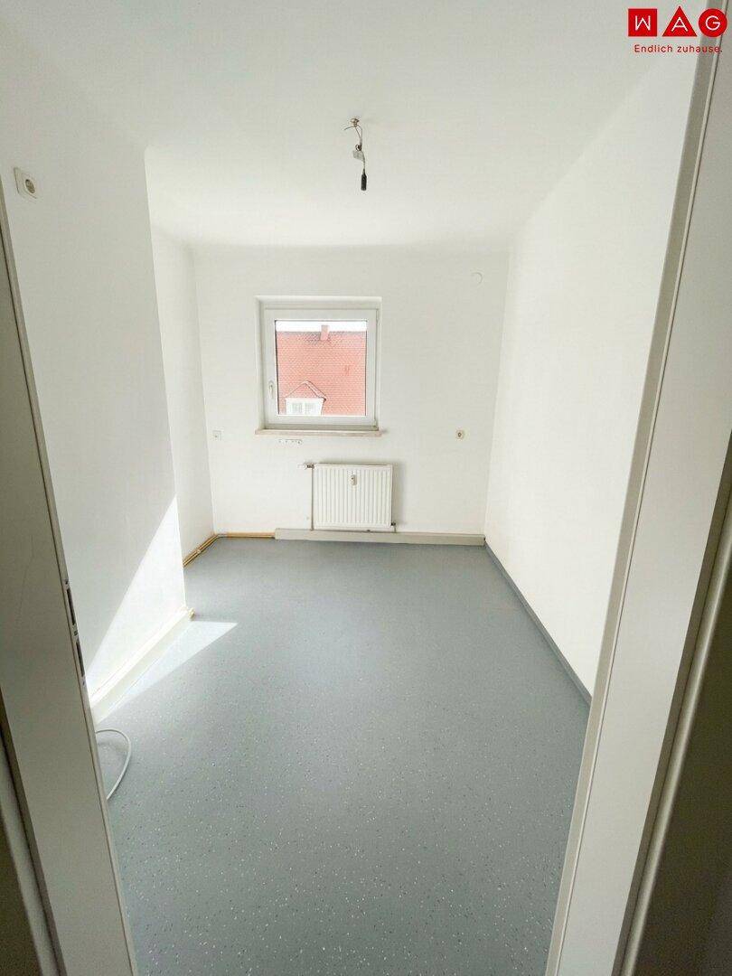 Leeres Zimmer mit grauem Vinylboden, weiß gestrichenen Wänden und einem Fenster.