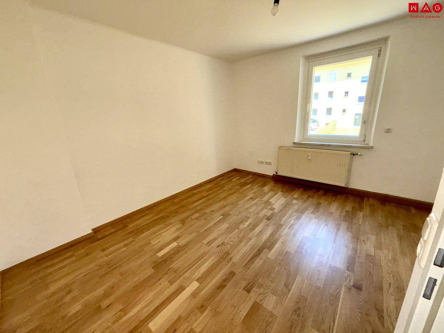 Leeres Zimmer mit hellem Parkettboden und einem Fenster mit Blick ins Grüne.