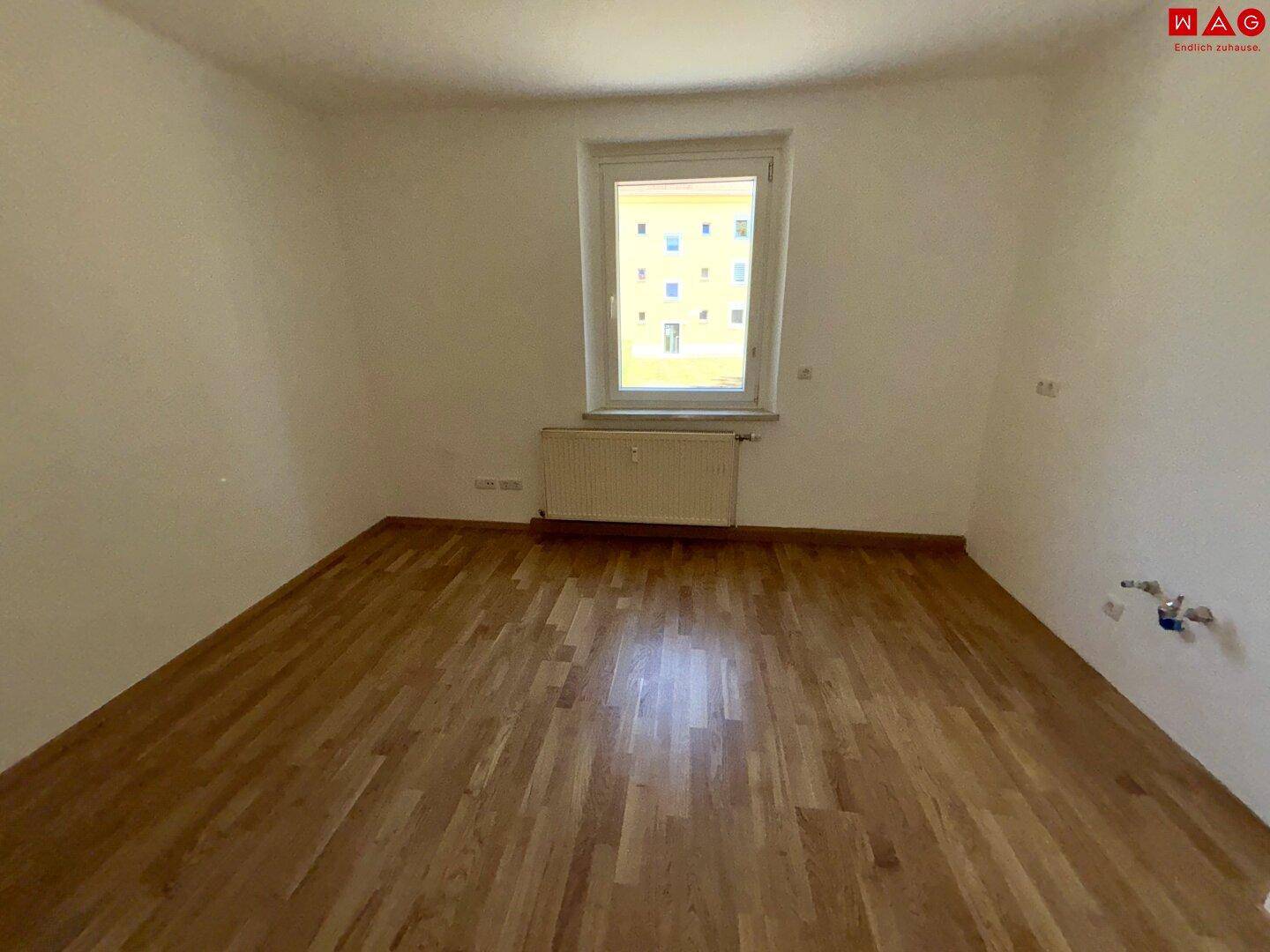 Freundliches Zimmer mit Parkettboden und Fenster, das viel Tageslicht hereinlässt.