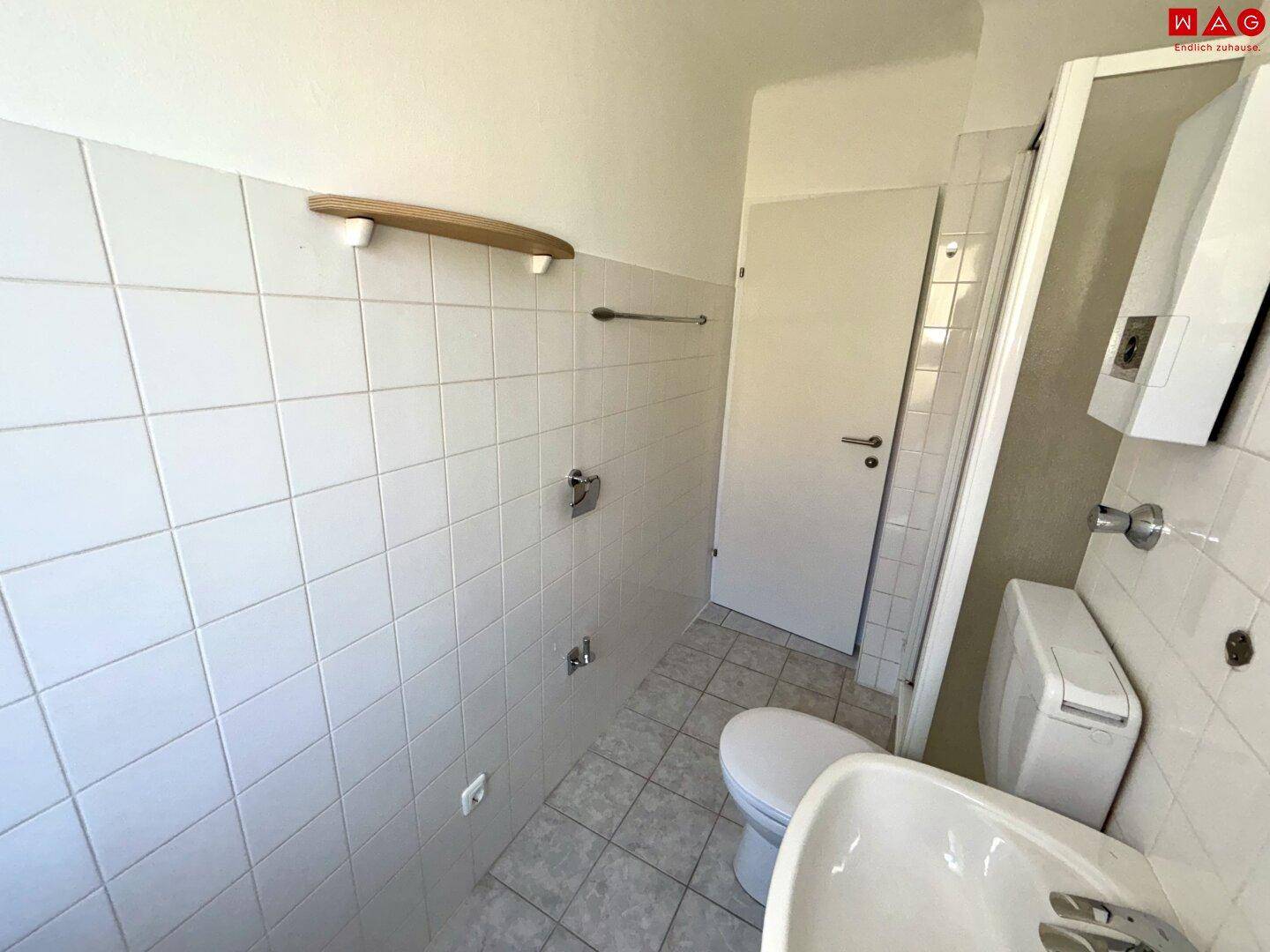 Einfaches Badezimmer mit weißen Fliesen, Waschbecken und Spiegel, sauber und gepflegt.