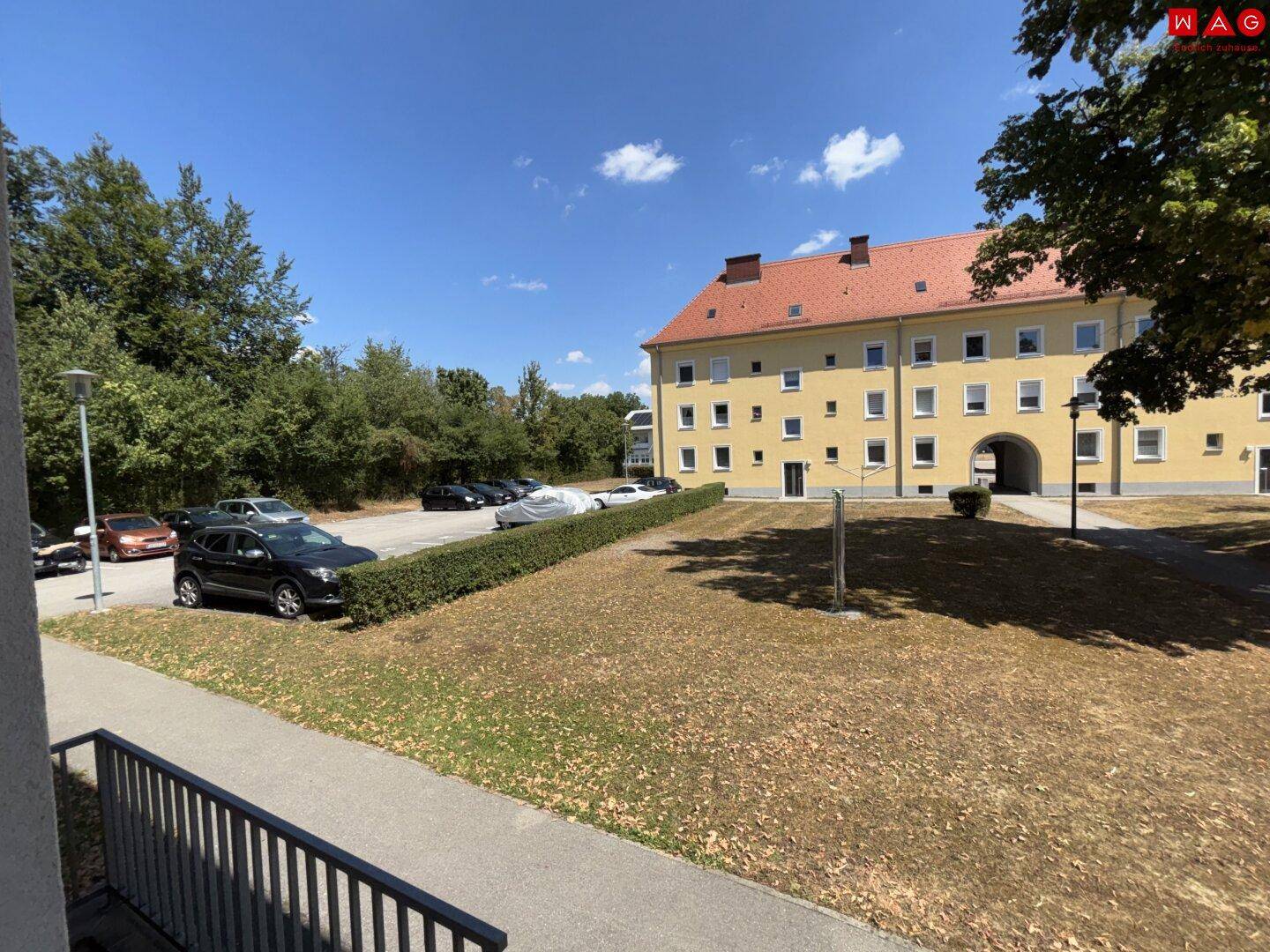 Wohngebäude mit Parkplätzen und Grünflächen unter blauem Himmel.