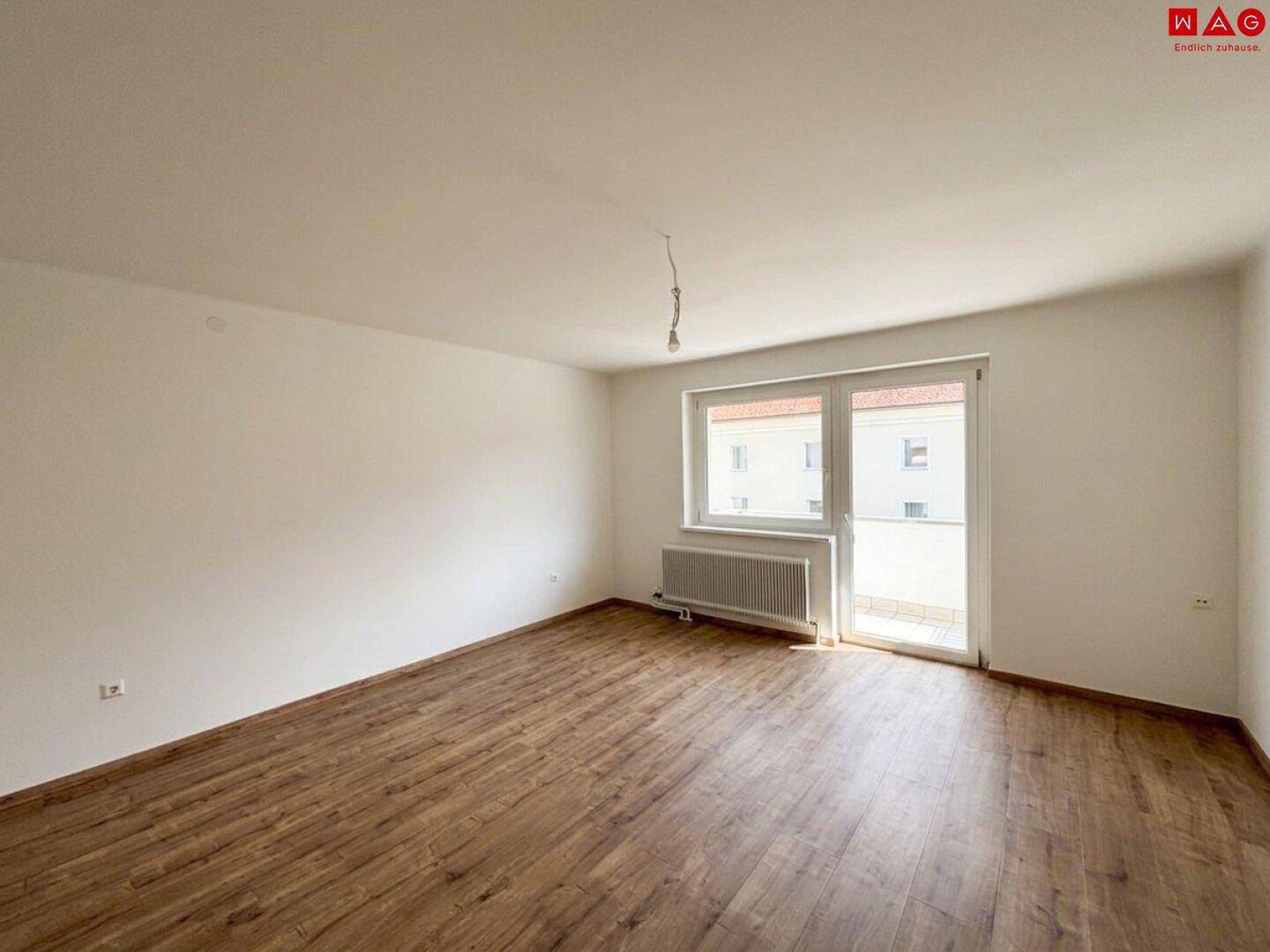 Geräumiges, leeres Zimmer mit Laminatboden, weißen Wänden und Zugang zu einem Balkon.