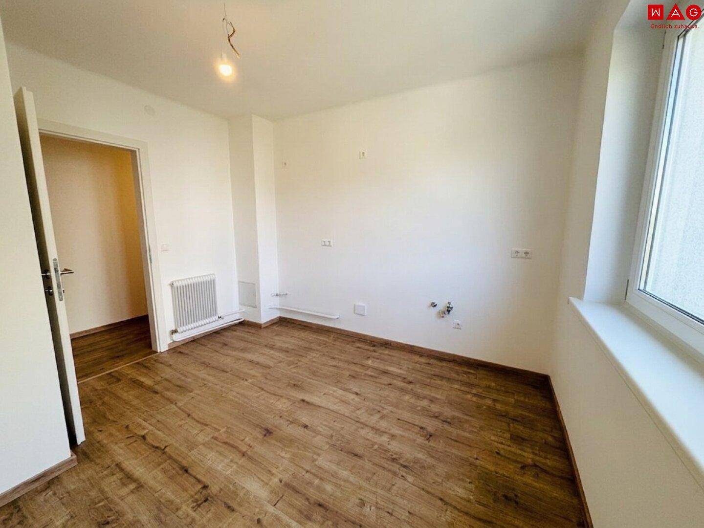 Leerer Raum mit Laminatboden, weißen Wänden und sichtbaren Anschlüssen für eine mögliche Küche.