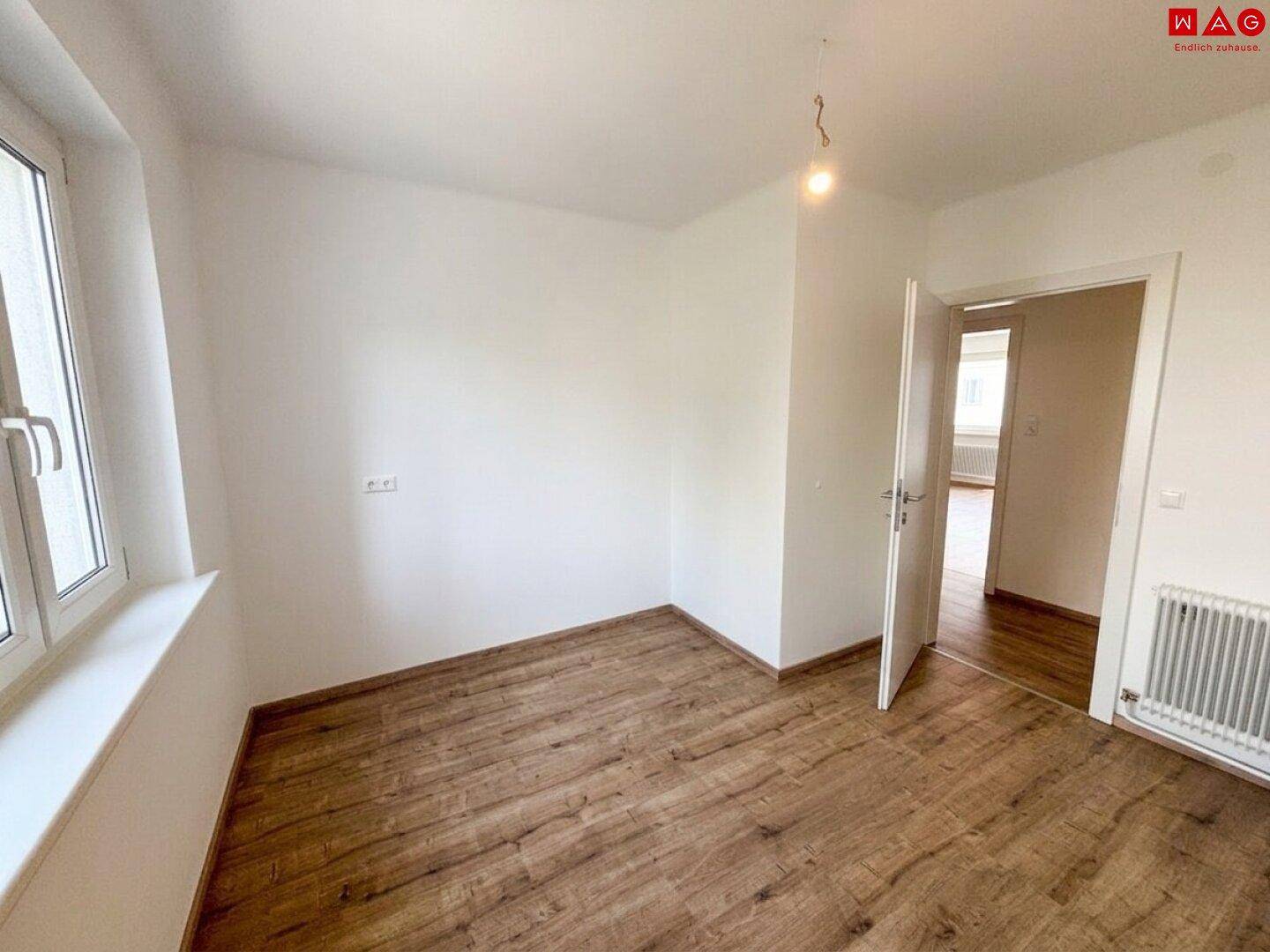 Helles, leeres Zimmer mit Laminatboden, weißen Wänden und offener Tür zu einem weiteren Raum.