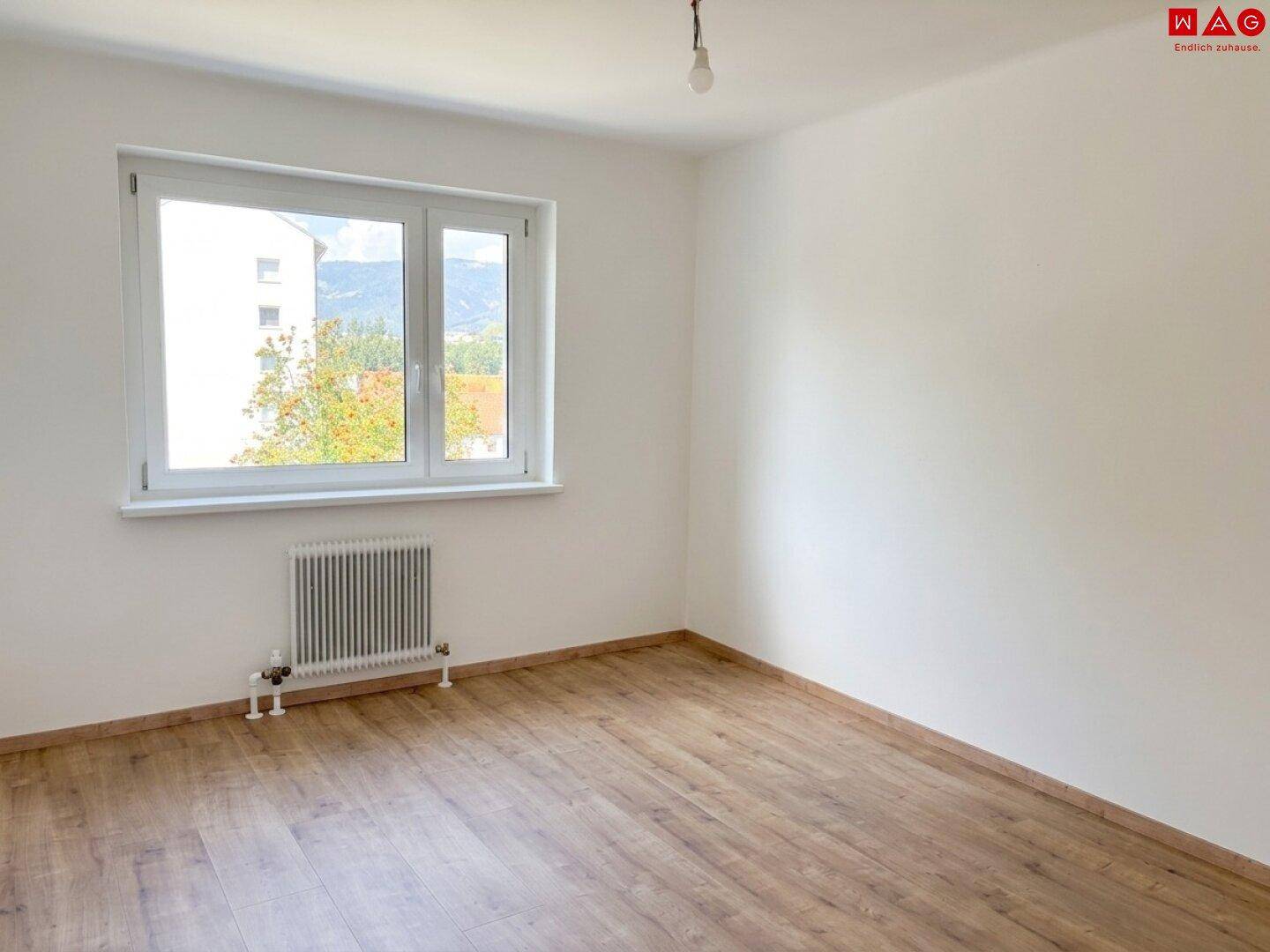Leeres Zimmer mit Laminatboden, Heizkörper und Fenster mit Fernblick auf die Berge.