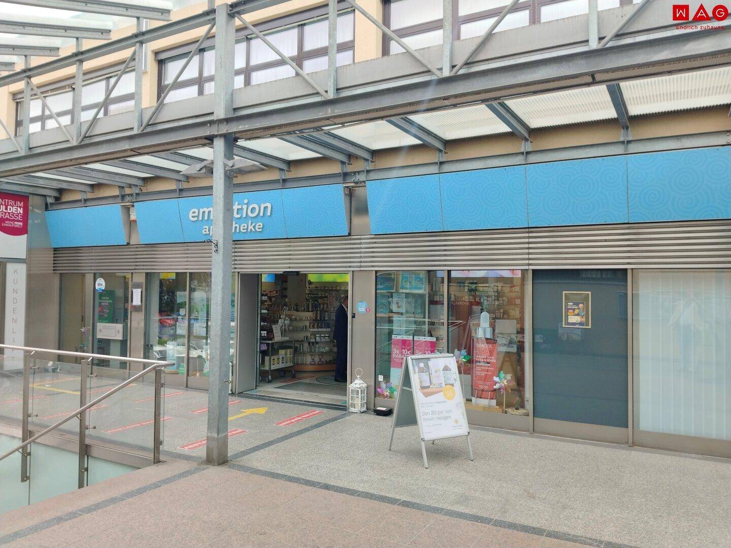 Breitere Ansicht des Apotheken-Eingangs mit blauen Schildern und einer zugänglichen Rampe.