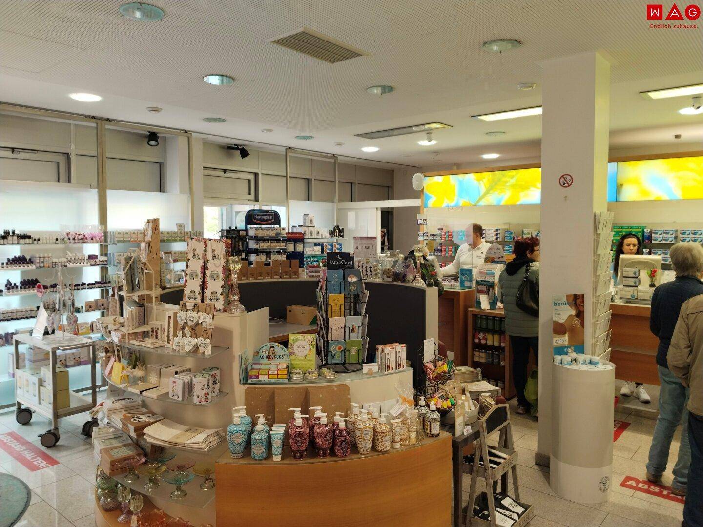 Hauptverkaufsbereich der Apotheke mit Theke, verschiedenen Produktpräsentationen und Kunden.