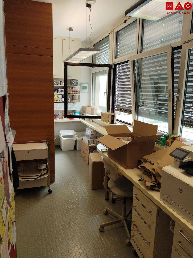 Hinterer Bürobereich oder Lagerraum mit Schreibtisch, Drucker und großen Fenstern mit Jalousien.