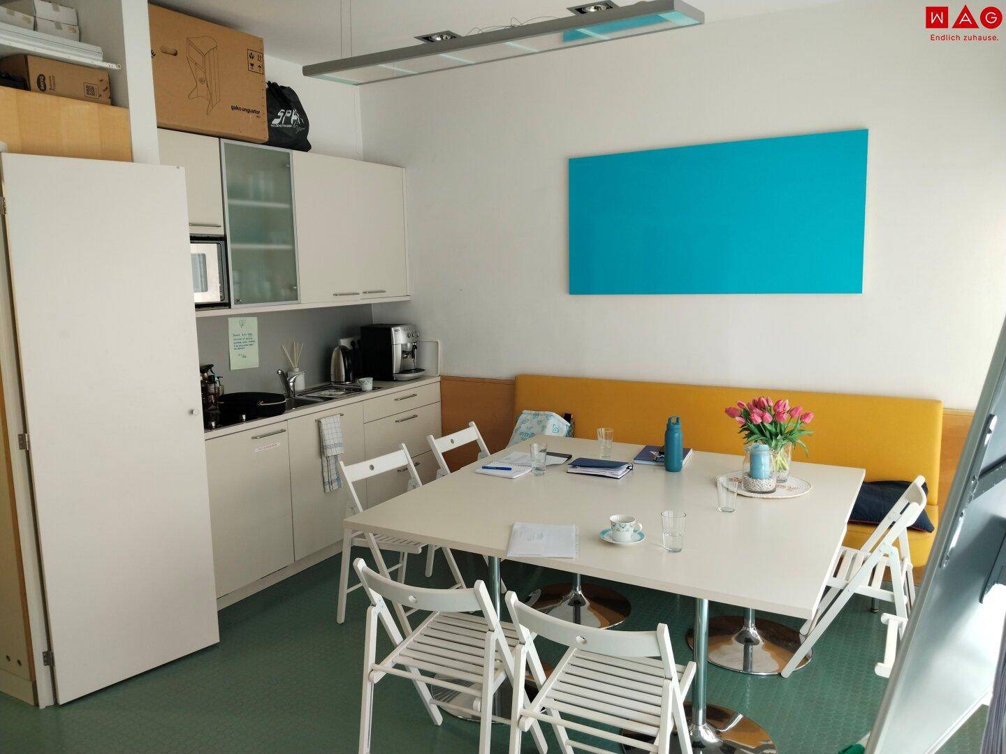 Pausen- oder Besprechungsraum mit kleiner Küchenzeile, großem Tisch und gelber Sitzbank.