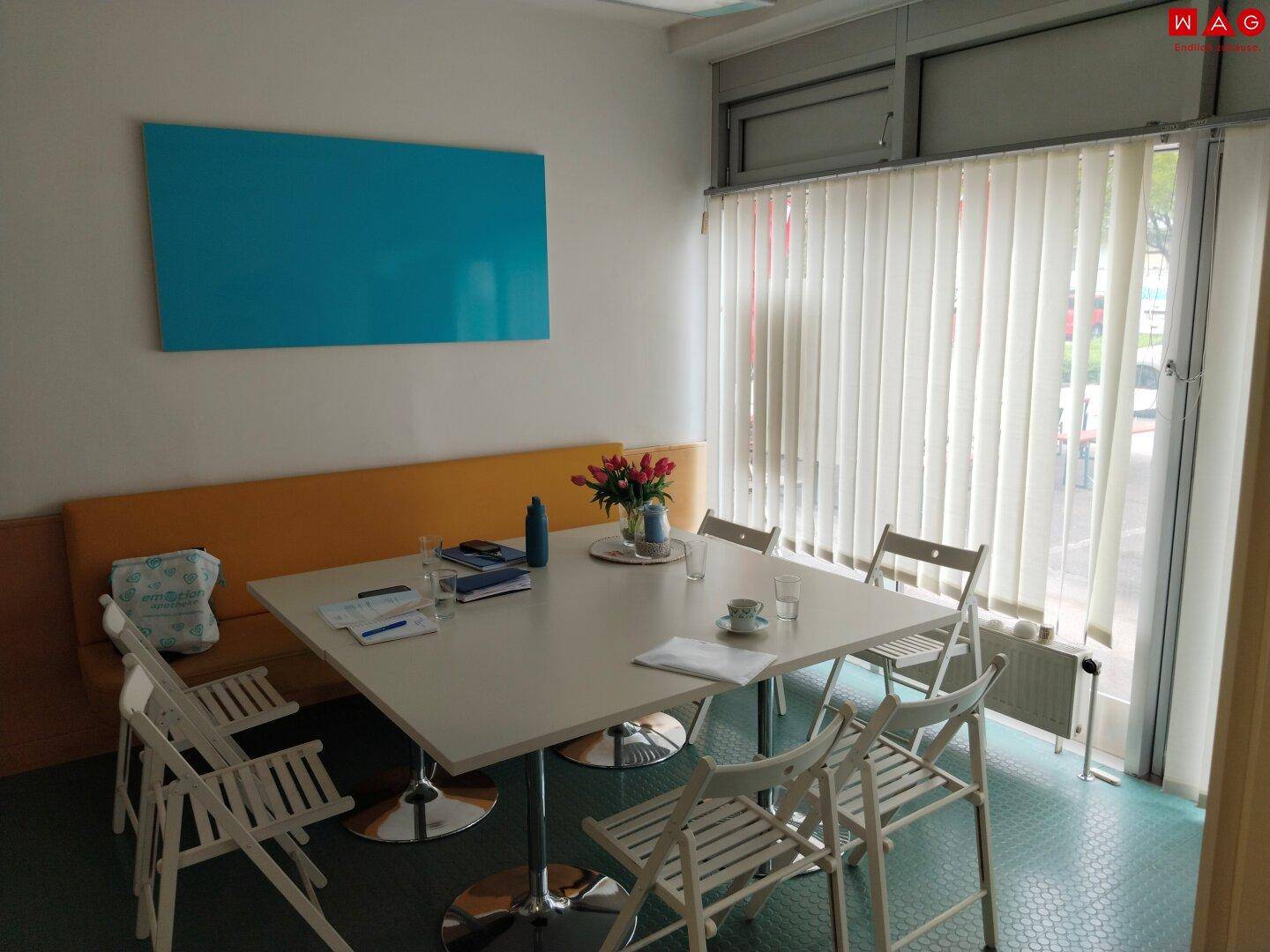 Pausenraum mit großem Tisch, Stühlen und Fenster mit vertikalen Jalousien für Privatsphäre.
