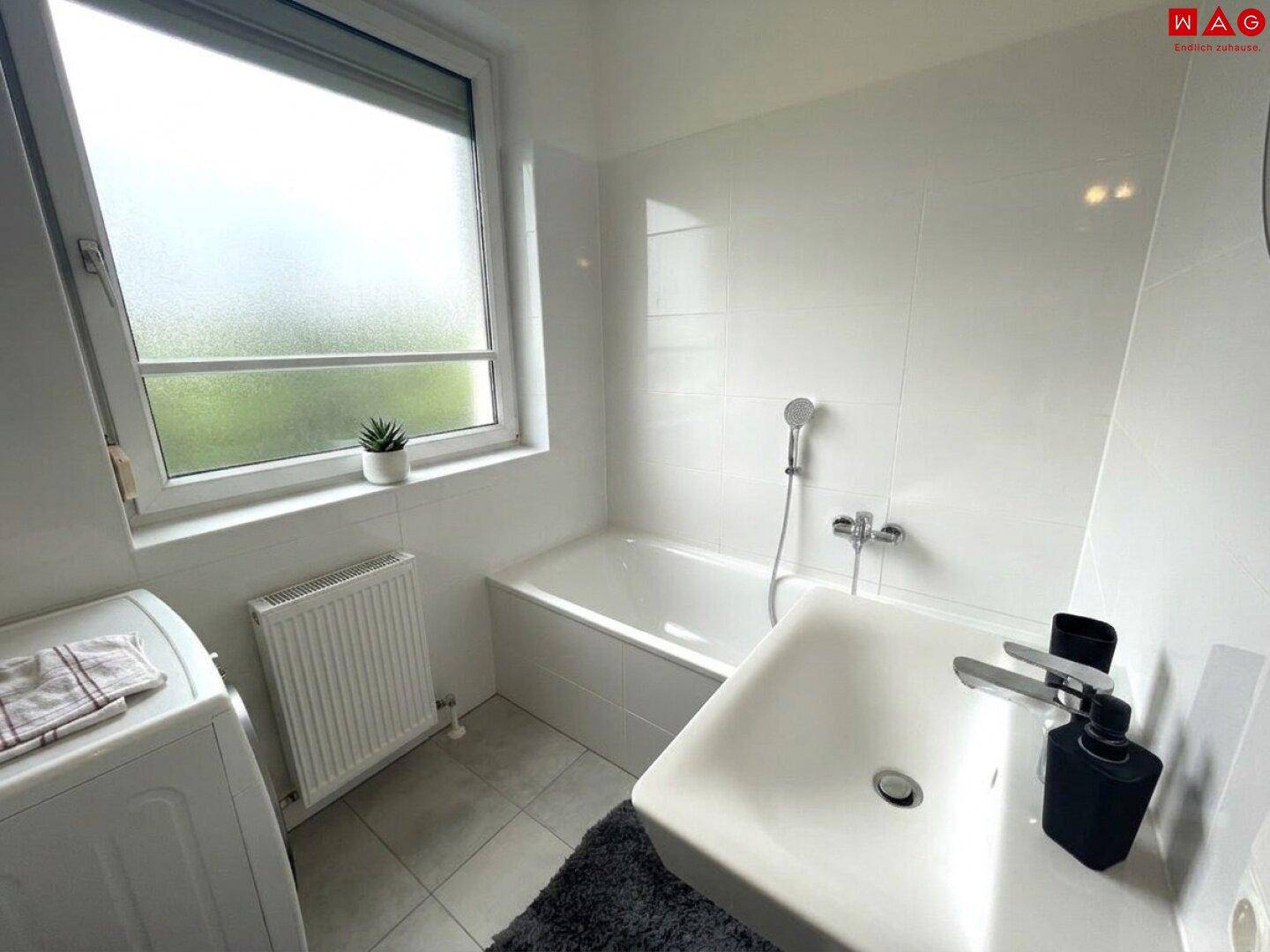 Helles Badezimmer mit Badewanne, Waschbecken, Waschmaschine und einem Fenster.