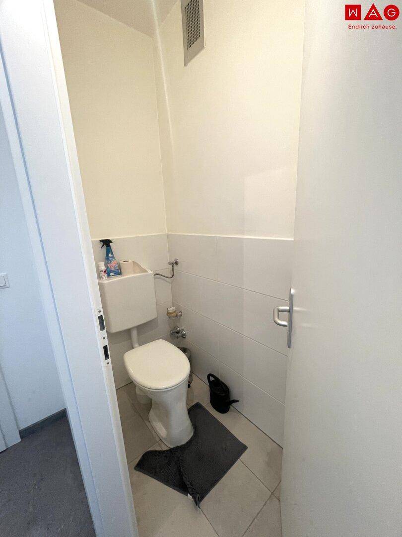 Separates WC mit Toilette, kleinem Waschbecken und weißen Fliesen an den Wänden.