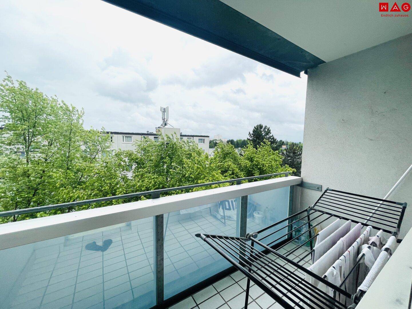 Balkon mit Wäscheständer, Fliesenboden und Blick auf Bäume und umliegende Gebäude.