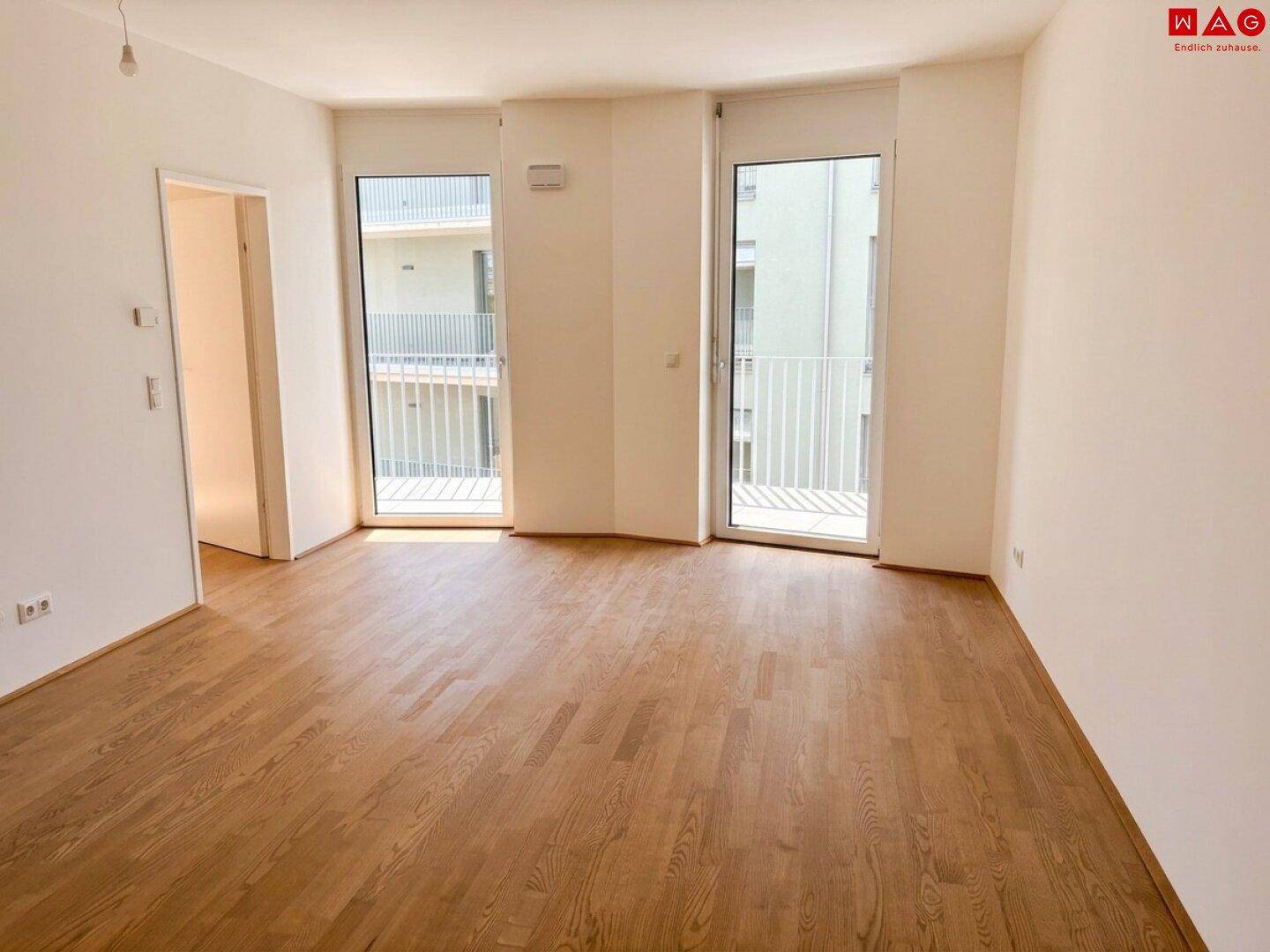 Geräumiges, unmöbliertes Wohnzimmer mit Holzboden und zwei Balkonzugängen, sehr hell.