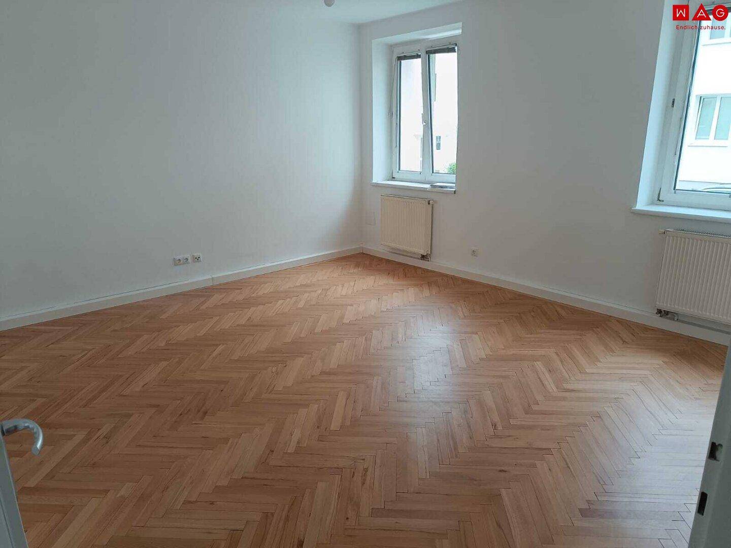 Geräumiges Zimmer mit hellem Fischgrätparkett, zwei Fenstern und weißen Wänden, ideal als Schlaf- oder Wohnbereich.