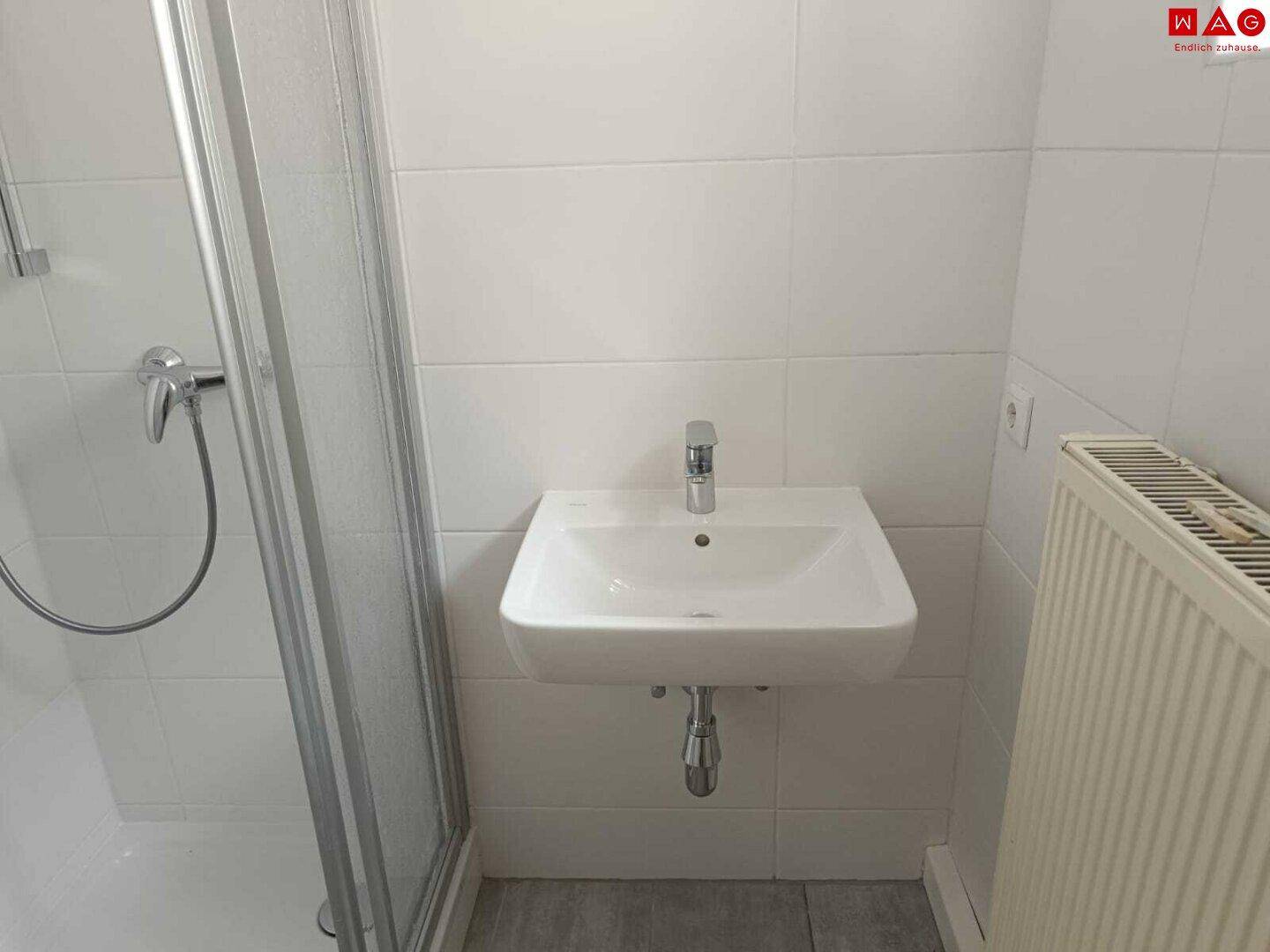 Modernes Badezimmer mit weißen Fliesen, wandhängendem Waschbecken, Duschkabine und Heizkörper.
