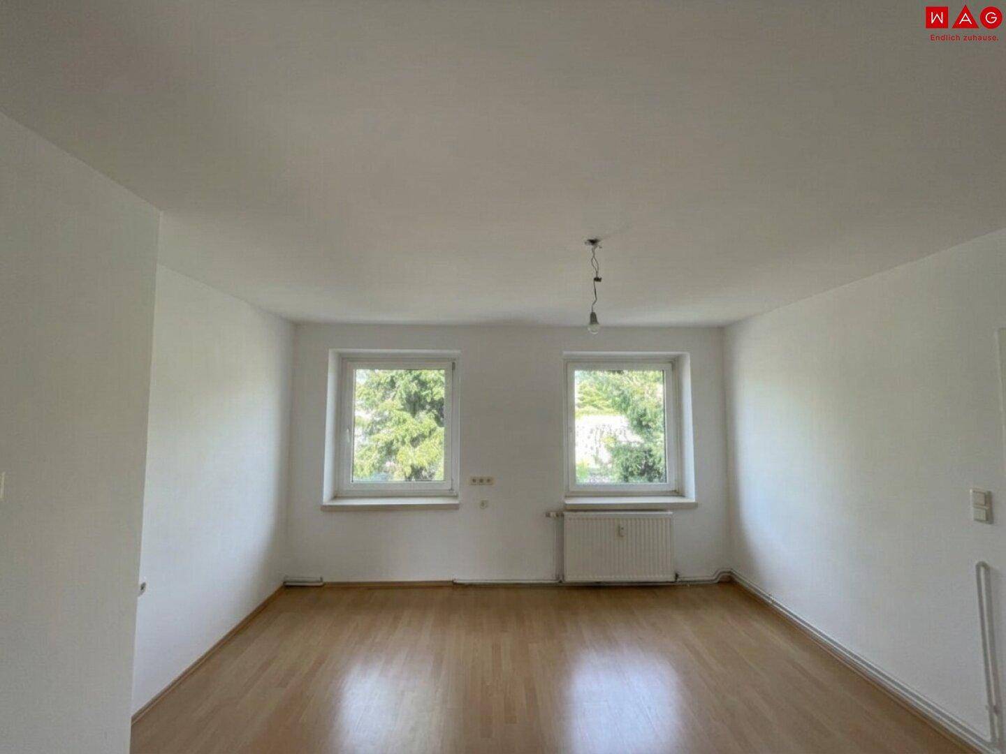 Lichtdurchfluteter Wohnraum mit zwei Fenstern, Heizkörper und hellem Laminatboden.
