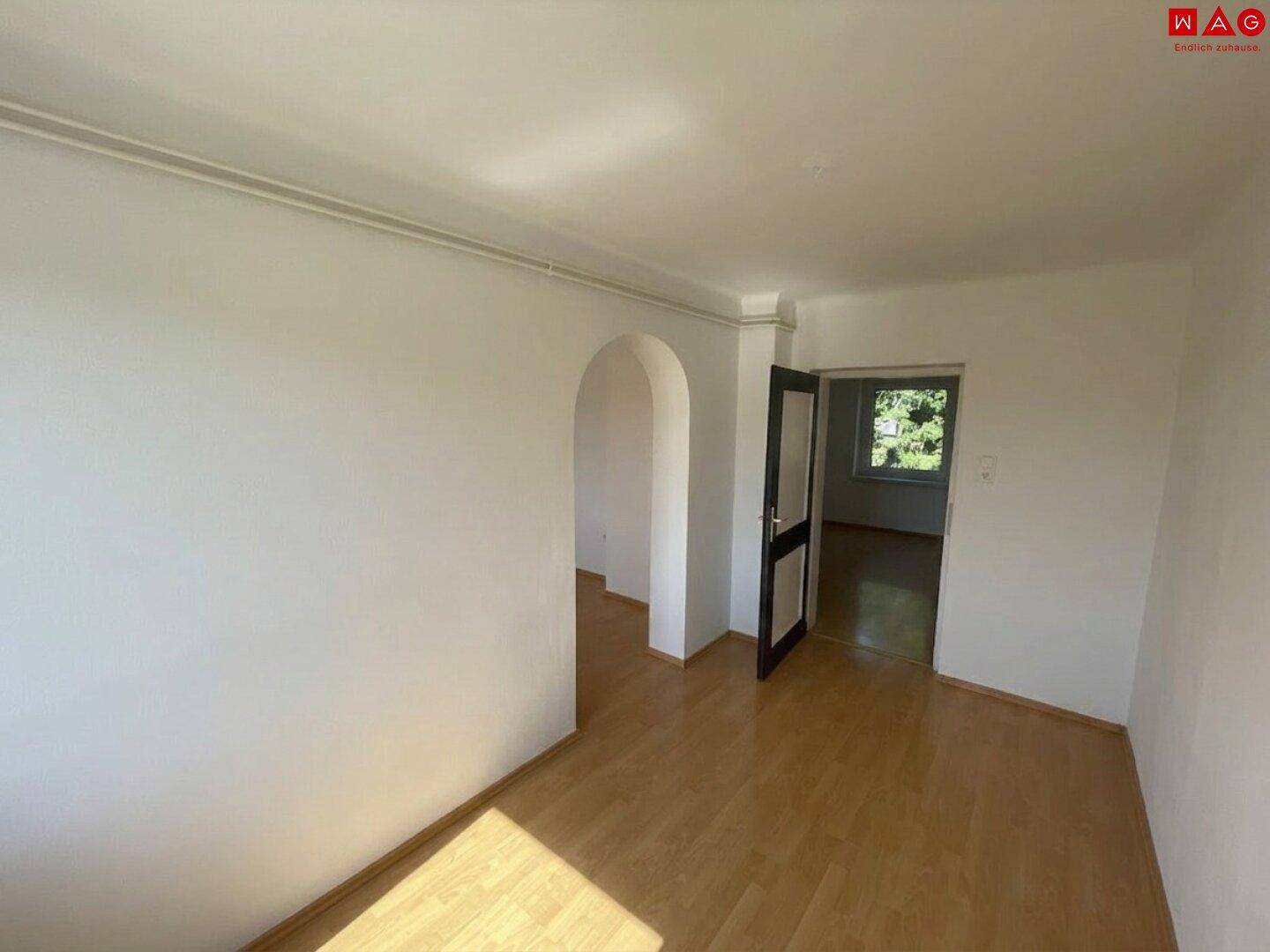 Wohnraum mit hellem Laminatboden, einem Rundbogen und Zugang zu weiteren Zimmern.
