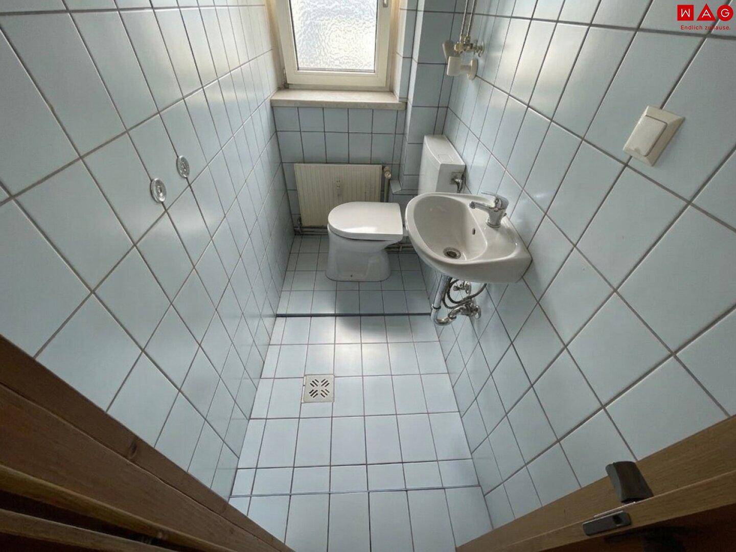 Kleines WC mit hellblauen Fliesen, Waschbecken, Toilette und Fenster.