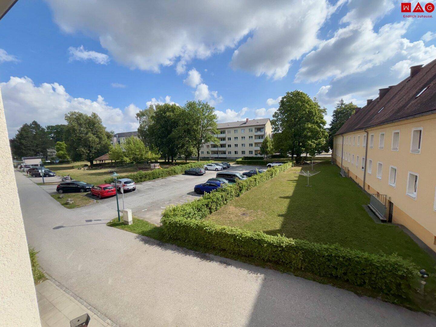 Blick auf den Parkplatz, Grünflächen und umliegende Wohngebäude unter blauem Himmel.