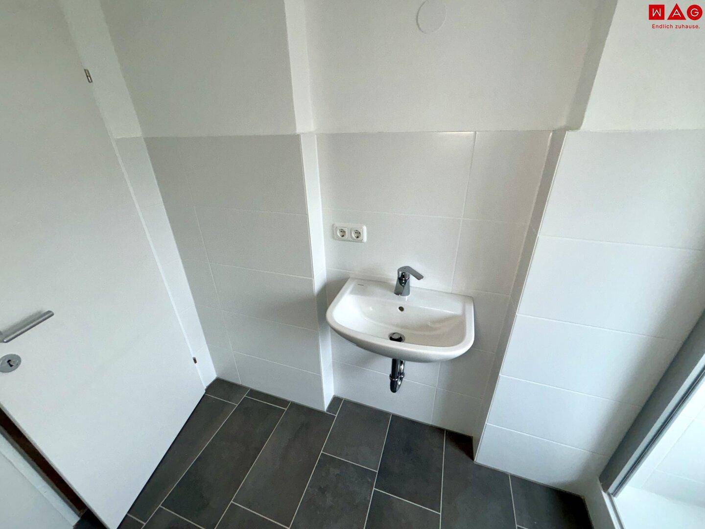 Wandmontiertes Waschbecken in einem hell gefliesten Badezimmer mit Steckdose.