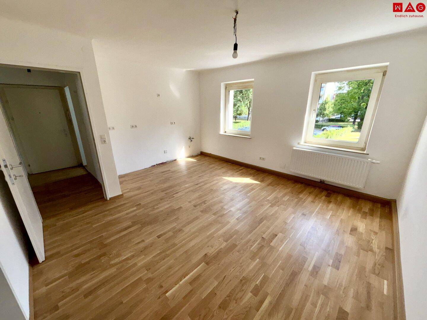 Geräumiges, leeres Zimmer mit hellem Holzparkettboden und zwei Fenstern, ideal als Wohnzimmer.