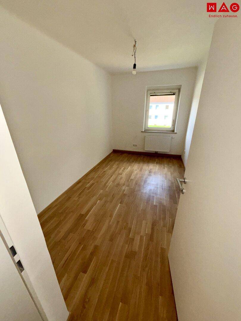 Leeres Schlafzimmer mit Parkettboden und Fenster, das viel Tageslicht hereinlässt.
