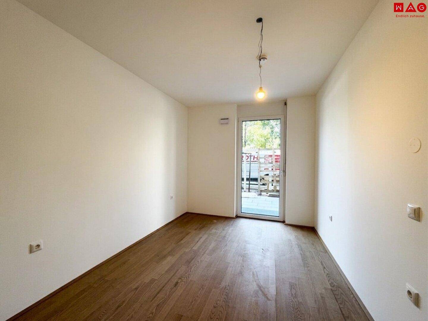 Heller Raum mit Holzboden und Zugang zu einem Balkon oder einer kleinen Terrasse.
