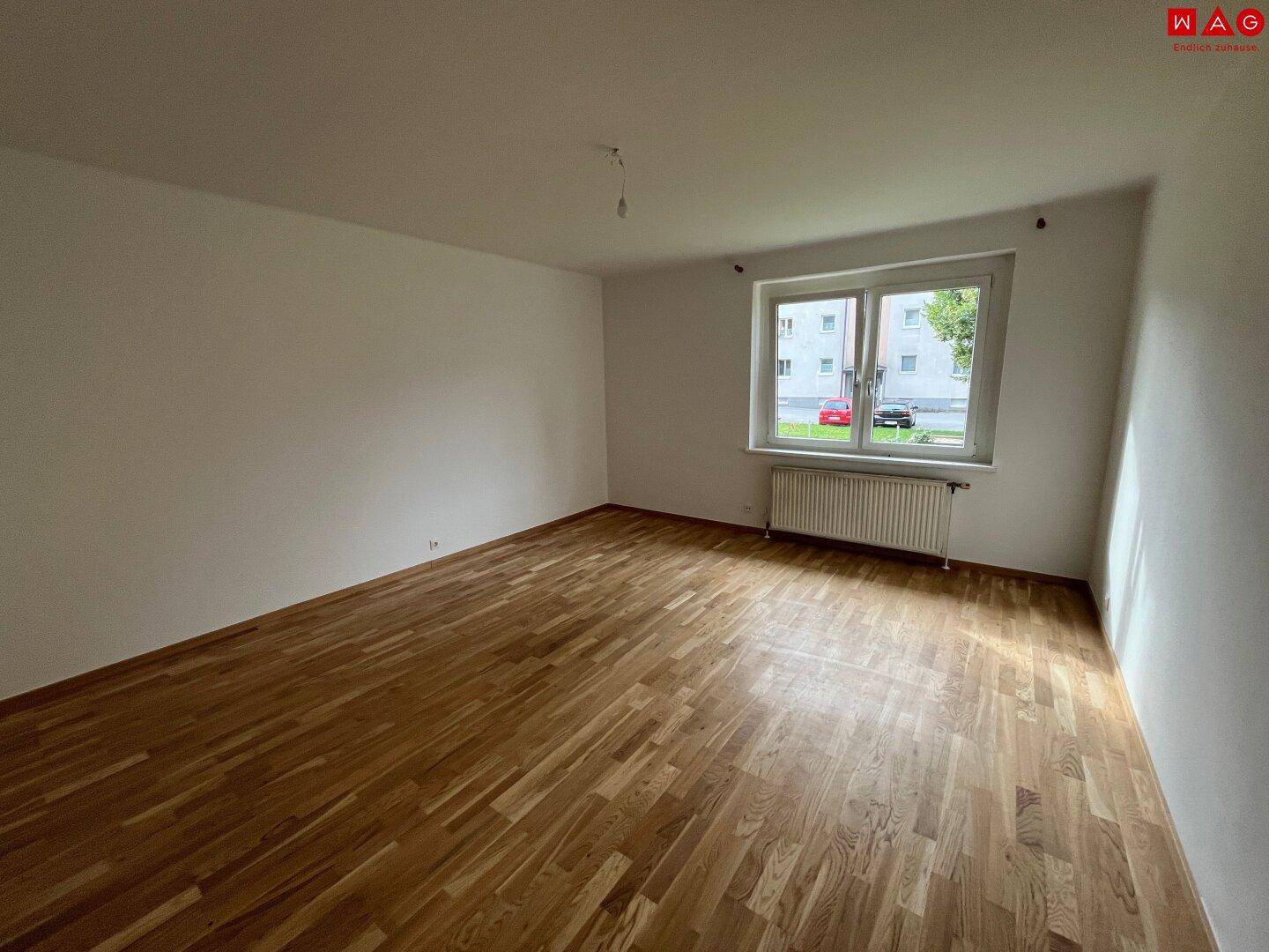 Heller, leerer Raum mit Holzboden und großem Fenster, ideal für Schlaf- oder Wohnzimmer.