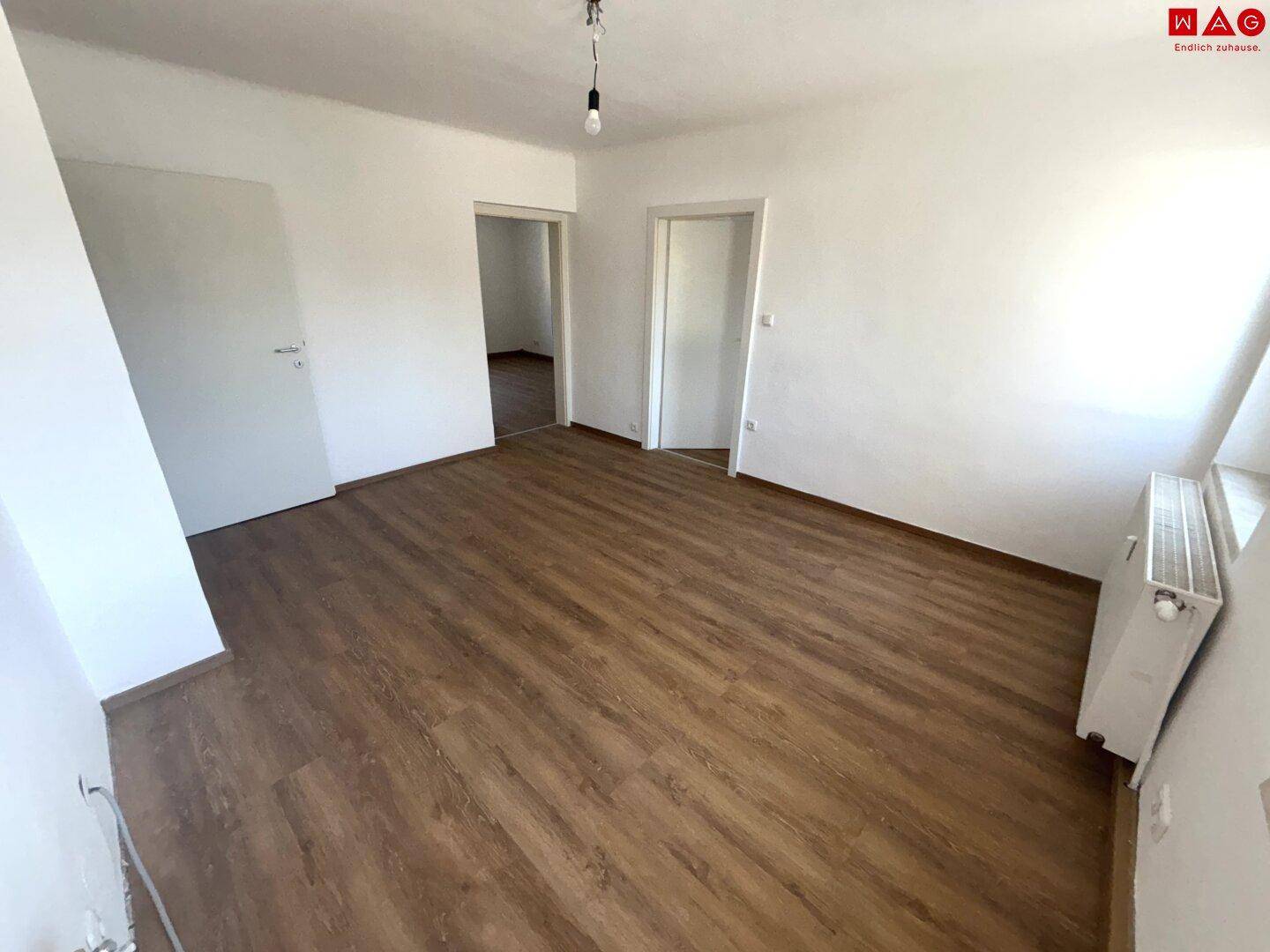 Helles Zimmer mit Laminatboden und Heizkörper, das durch mehrere Türen zugänglich ist.