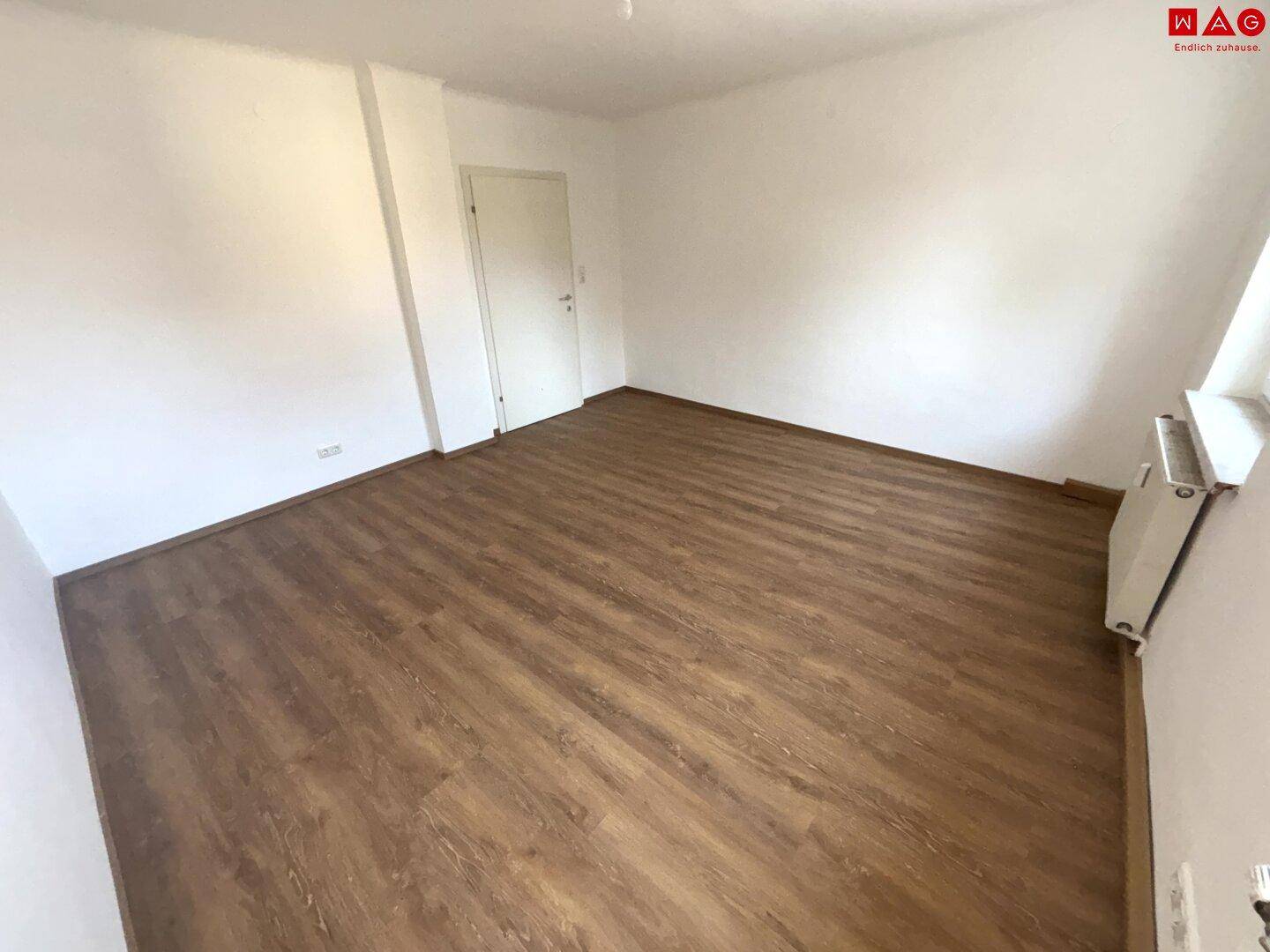 Leeres Zimmer mit Laminatboden und weißer Tür, das vielseitig genutzt werden kann.