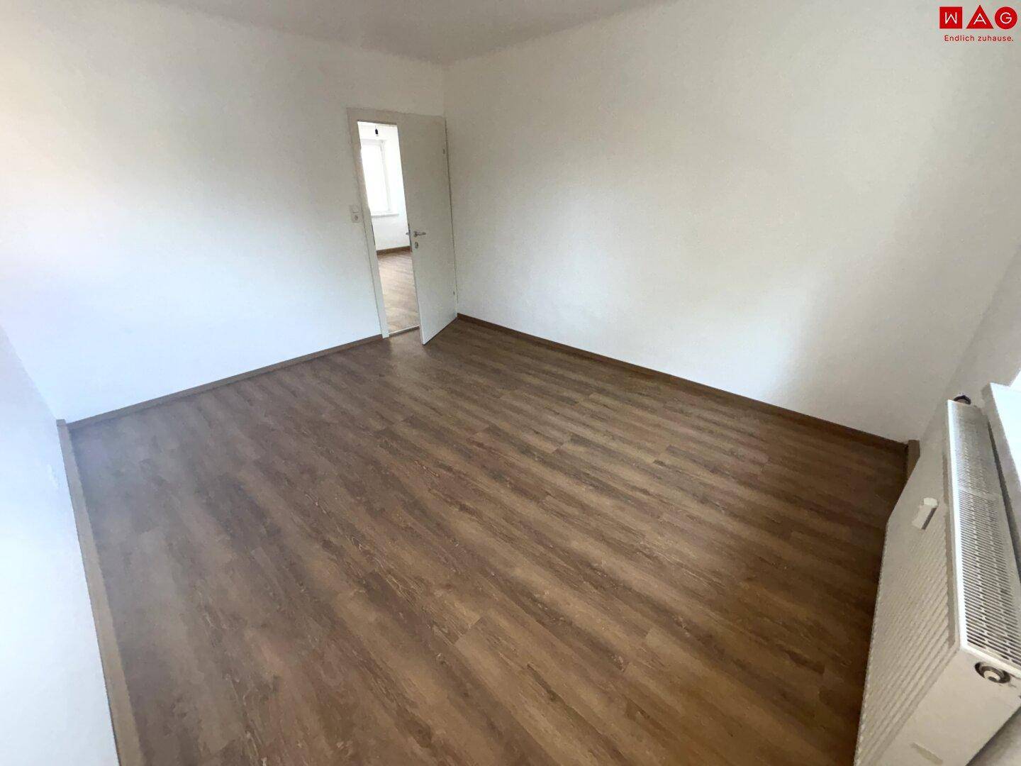 Helles Zimmer mit Laminatboden und weißer Tür, das einen direkten Zugang zu einem weiteren Raum ermöglicht.