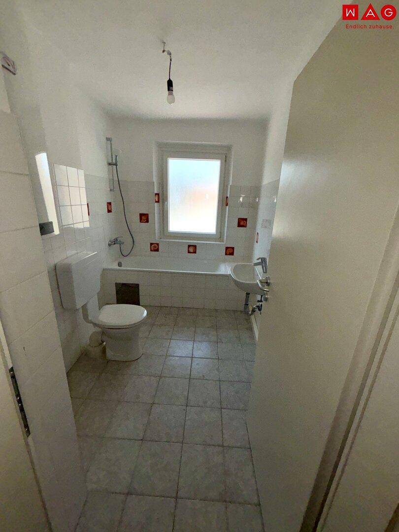 Badezimmer mit Badewanne, WC und Fenster, gefliest in Weiß mit roten Akzenten.