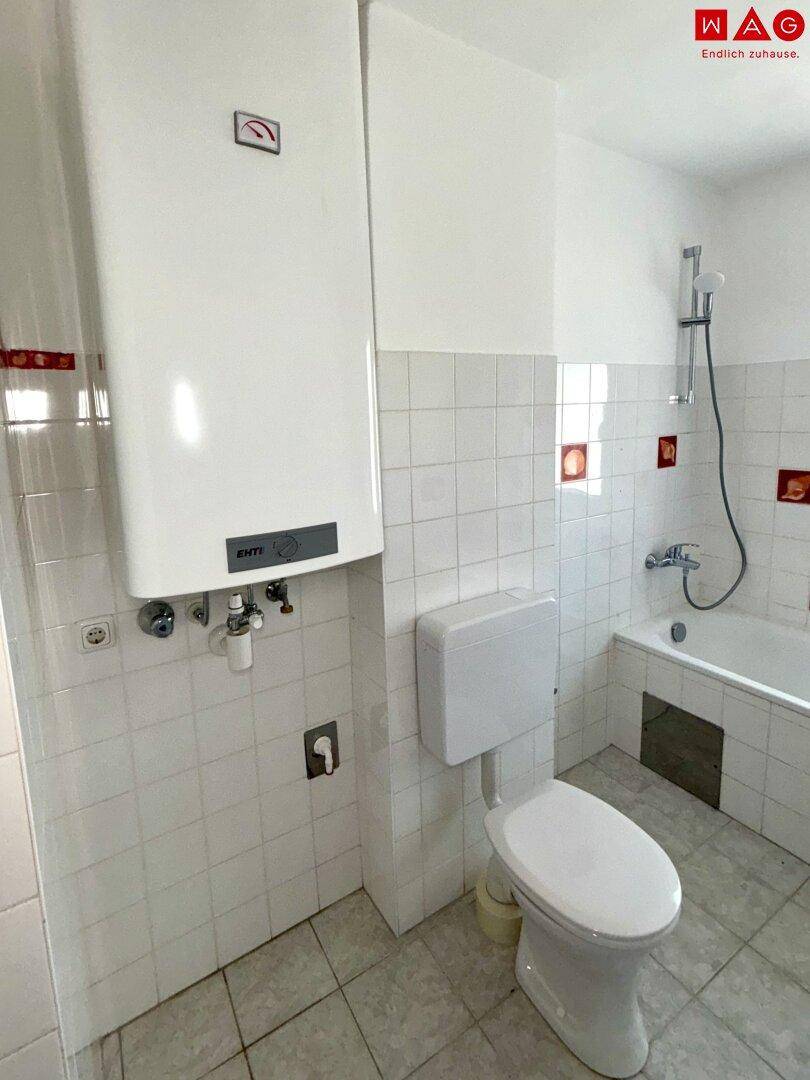Badezimmer mit Boiler, WC und Badewanne, gefliest in Weiß mit roten Dekorfliesen.