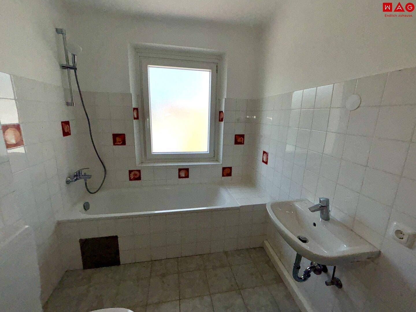 Badezimmer mit Badewanne, Waschbecken und Fenster, gefliest in Weiß mit roten Akzenten.