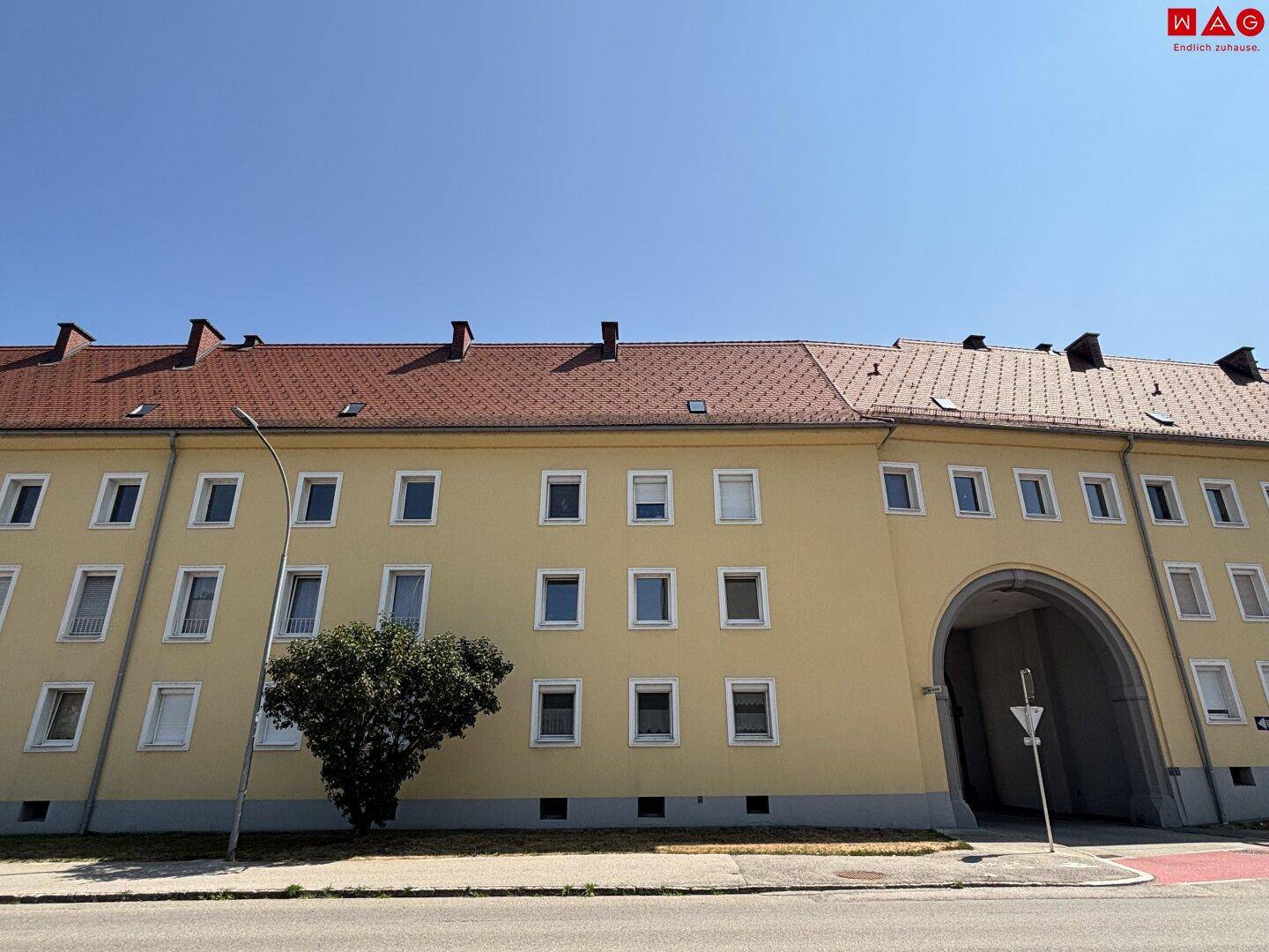 Gelbes Mehrfamilienhaus mit rotem Ziegeldach und einem großen Torbogen, umgeben von Grünflächen.
