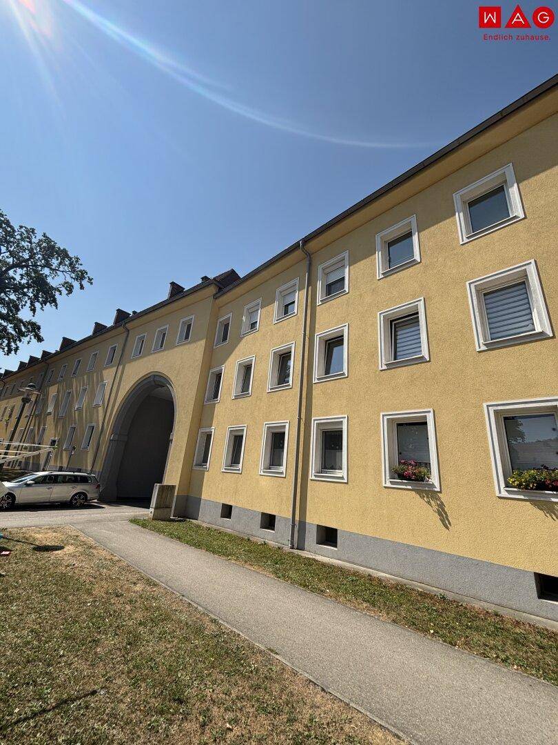 Gelbes Mehrfamilienhaus mit großem Torbogen und gepflegter Grünfläche vor dem Eingangsbereich.
