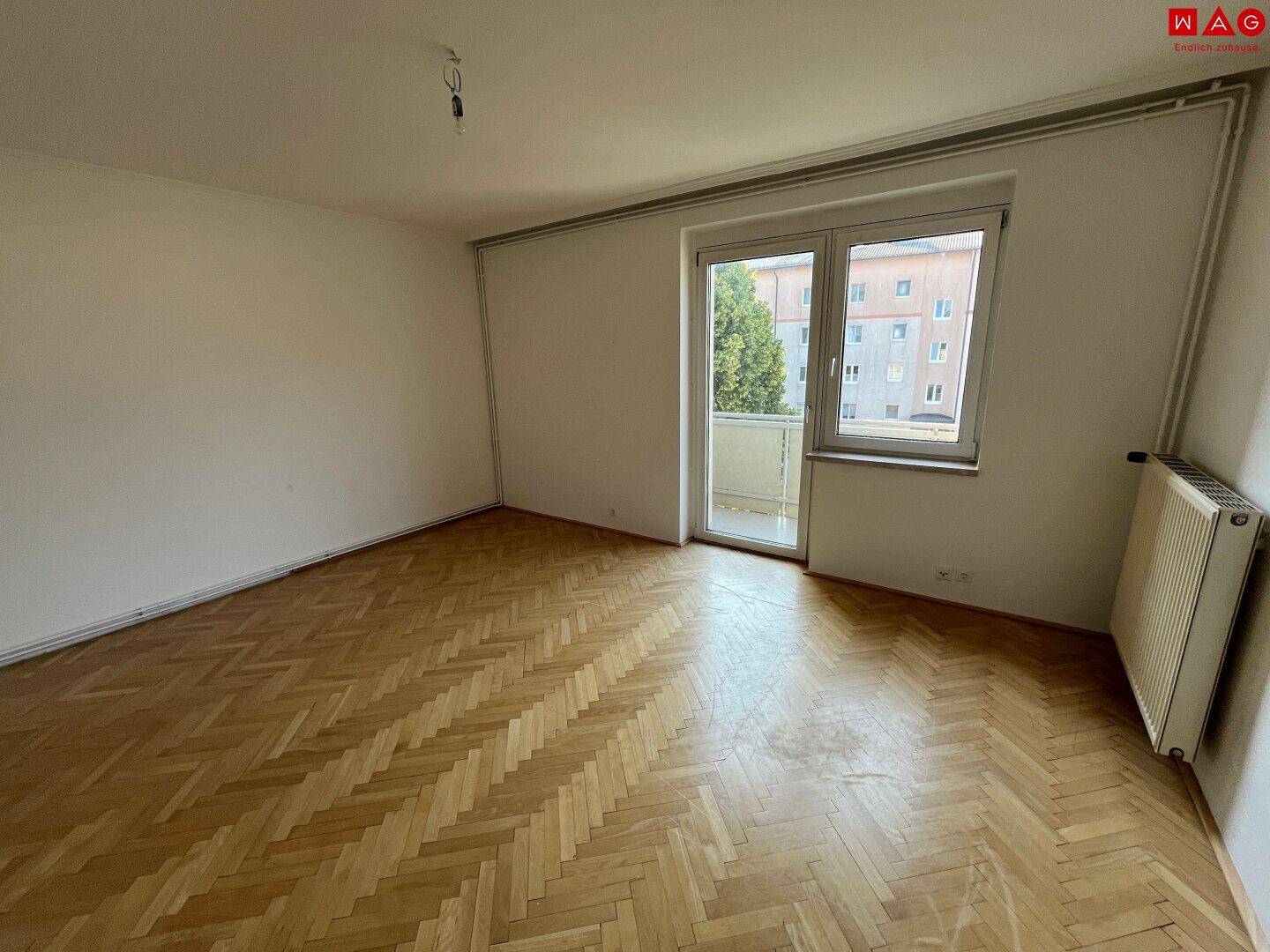 Leeres Zimmer mit Fischgrätparkettboden und Zugang zu einem Balkon.