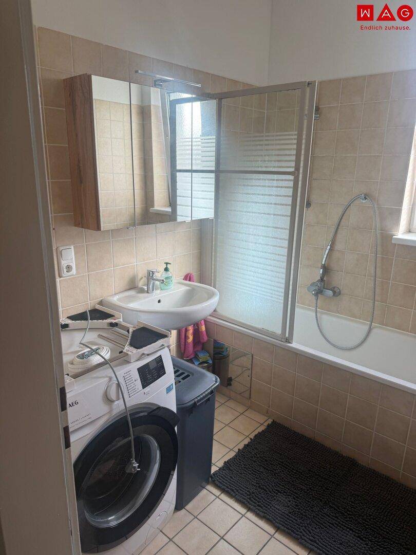 Badezimmer mit Badewanne, Waschbecken, Spiegelschrank und beigen Fliesen im 80er-Jahre-Stil.