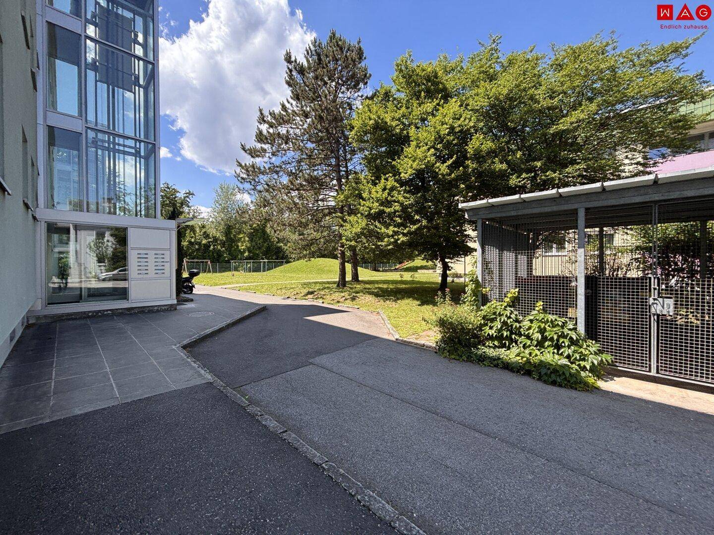 Außenbereich mit Glasaufzug, gepflastertem Weg und umzäuntem Abstellbereich neben Grünfläche.