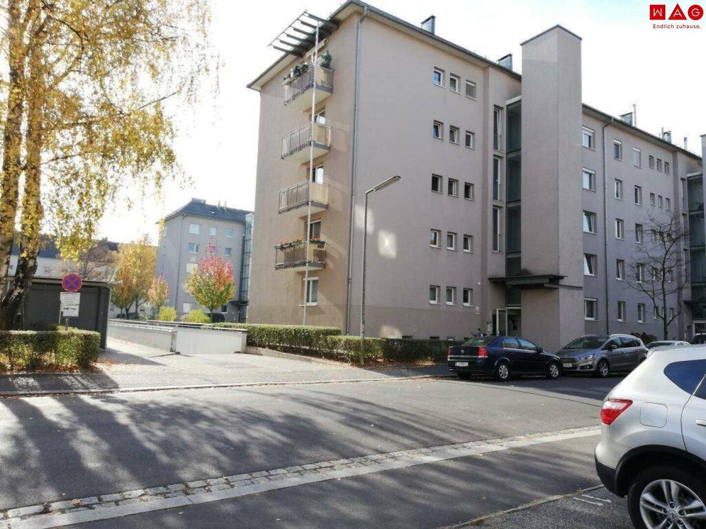 Mehrstöckiges Wohngebäude mit Balkonen und geparkten Autos auf der Straße, in einer städtischen Umgebung.